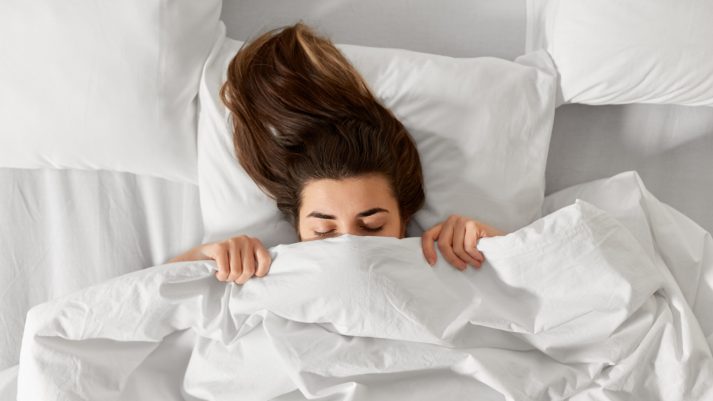 Abgebildet ist eine Frau die über die Bettdecke herausschaut und sich auch sicherlich die Frage gestellt: Ist nackt schlafen gesund?