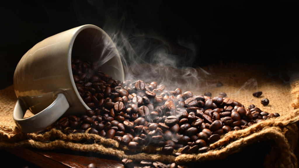 Abgebildet ist eine Kaffeetasse, in der aber kein flüssiger Kaffee enthalten ist, sondern warme Kaffeebohnen, die auf einen Sack ausgeschüttet werden