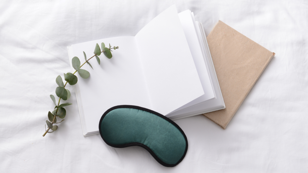 Abgebildet ist eine Schlafmaske die auf einer Bettdecke liegt, daneben sieht man ein offenes Buch.